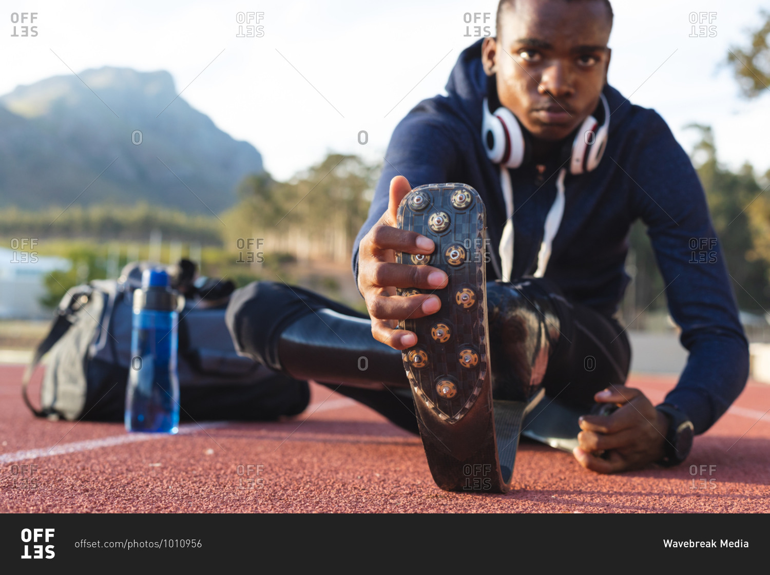Fit, mixed race disabled male athlete at an outdoor sports stadium, with gym bag and water bottle stretching on race track wearing running blades. Disability athletics sport training.