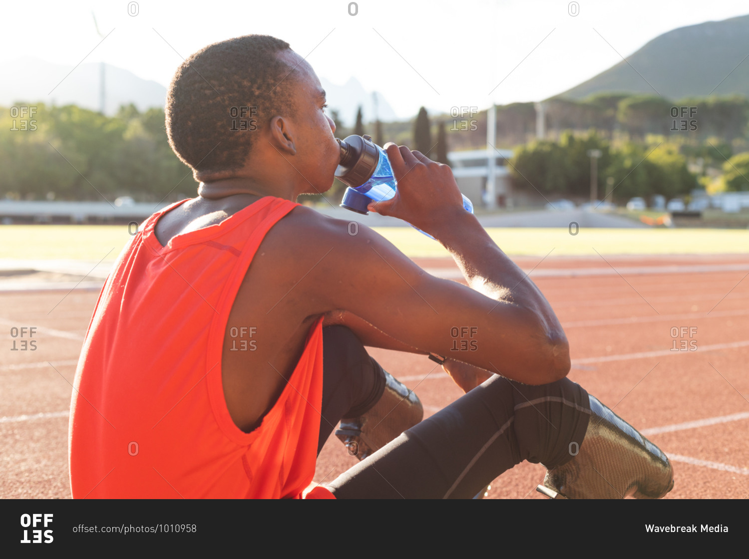 Fit, mixed race disabled male athlete at an outdoor sports stadium, resting and drinking from water bottle on race track wearing running blades. Disability athletics sport training.