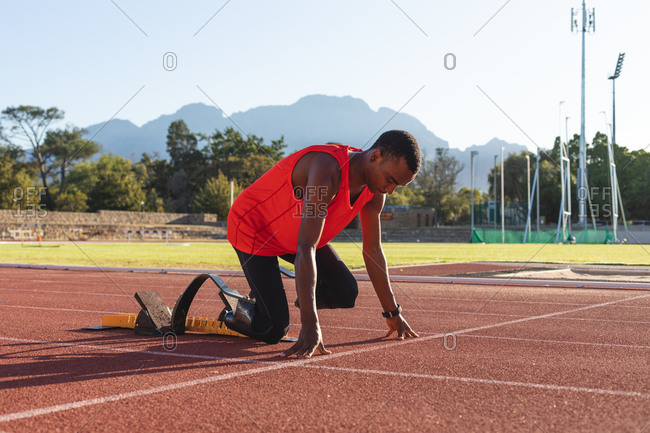 Fit, mixed race disabled male athlete at an outdoor sports stadium, kneeling in starting blocks on race track wearing running blades. Disability athletics sport training.