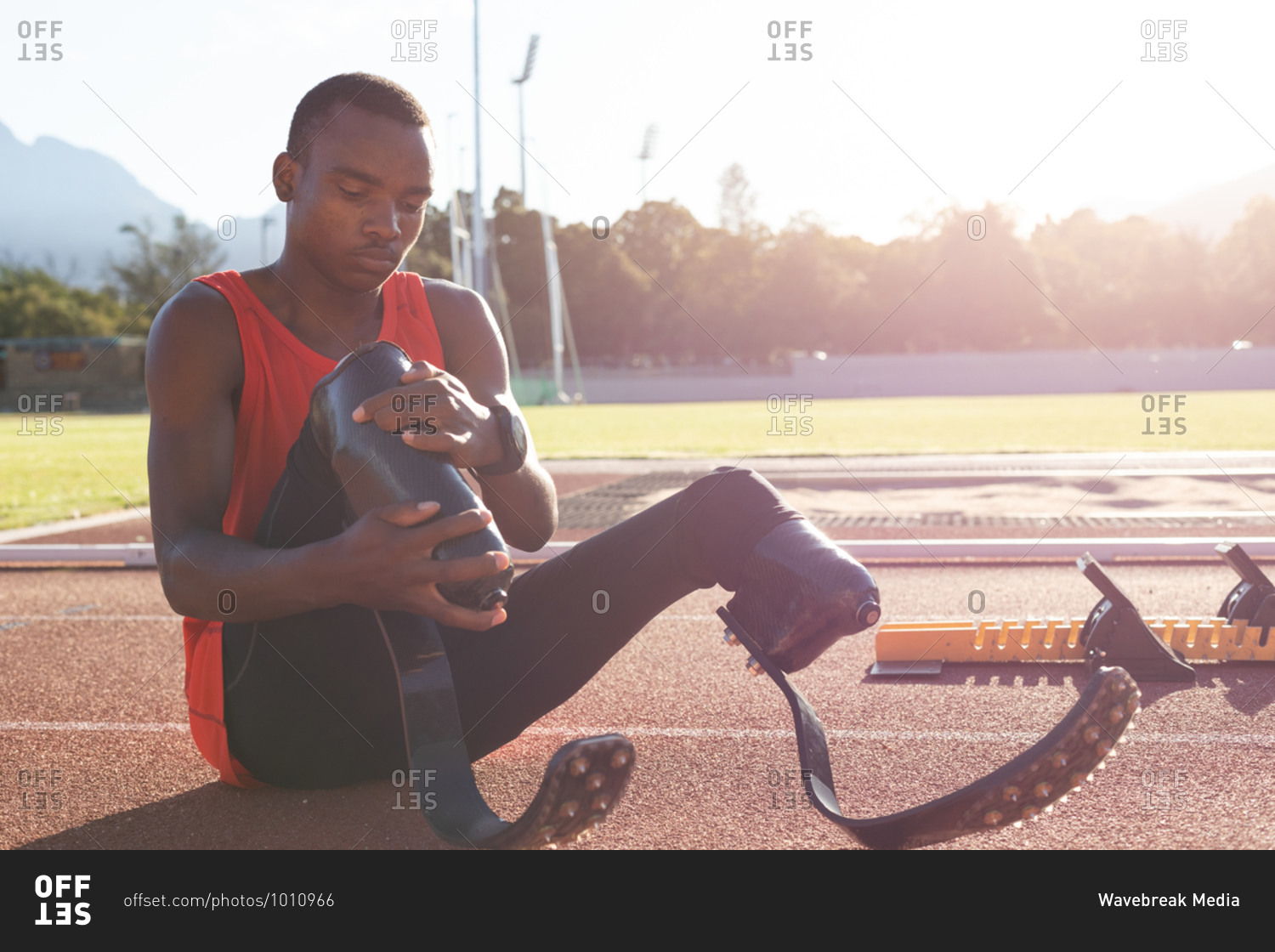 Fit, mixed race disabled male athlete at an outdoor sports stadium, sitting on race track adjusting running blades. Disability sport training.
