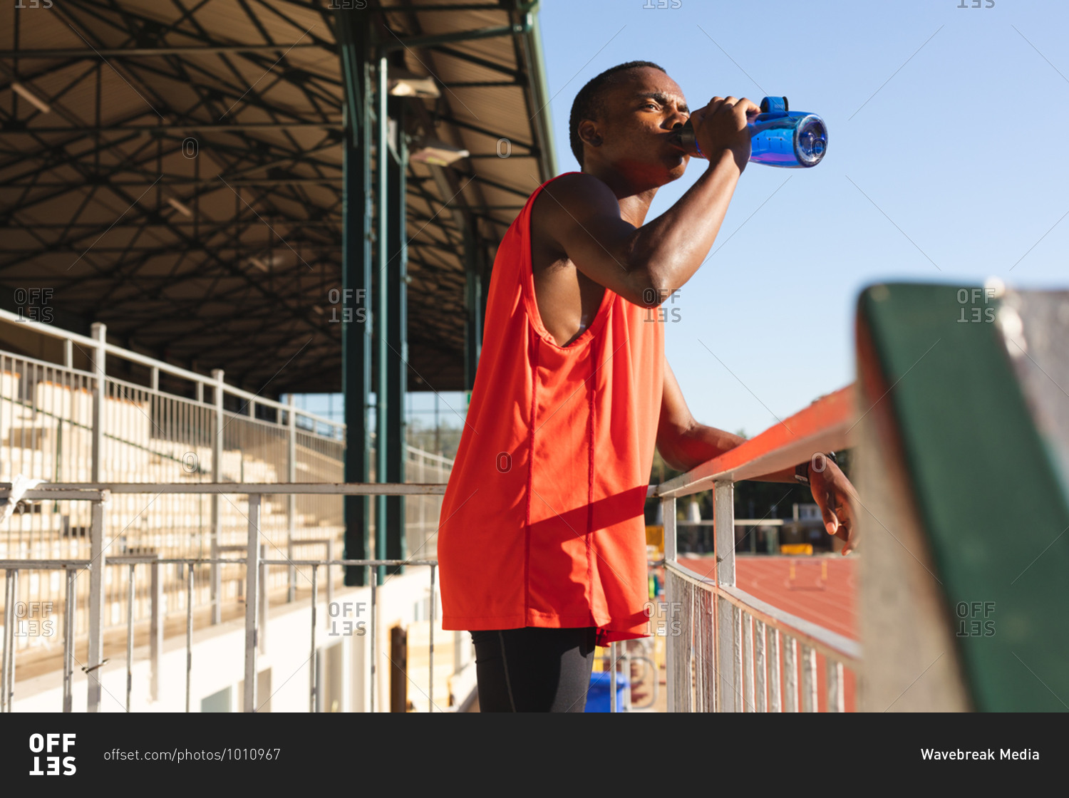 Fit, mixed race male athlete at an outdoor sports stadium, resting and drinking from water bottle standing in the stands. Athletics sport training.