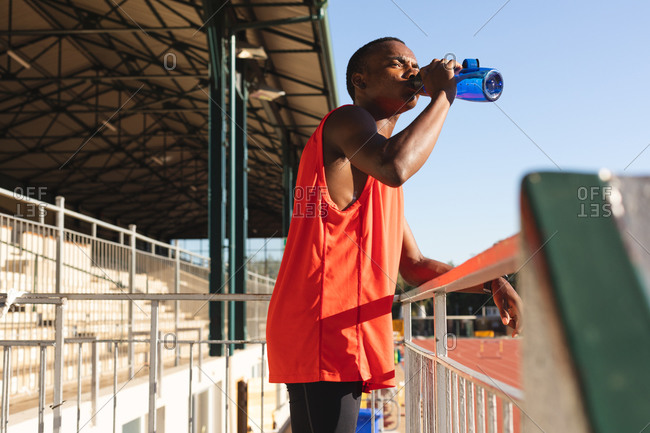 Fit, mixed race male athlete at an outdoor sports stadium, resting and drinking from water bottle standing in the stands. Athletics sport training.