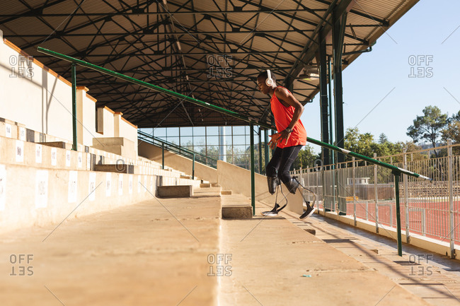 Fit, mixed race disabled male athlete at an outdoor sports stadium, walking up stairs in the stands wearing headphones and running blades. Disability athletics sport training.