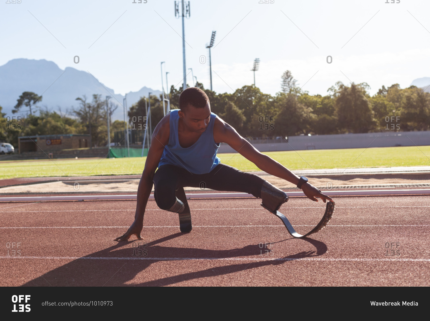 Fit, mixed race disabled male athlete at an outdoor sports stadium, preparing before workout stretching on race track wearing running blades. Disability athletics sport training.