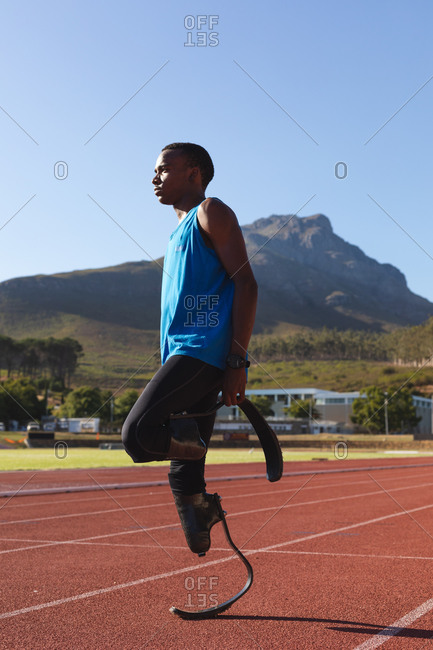Fit, mixed race disabled male athlete at an outdoor sports stadium, preparing before workout stretching on race track wearing running blades. Disability athletics sport training.
