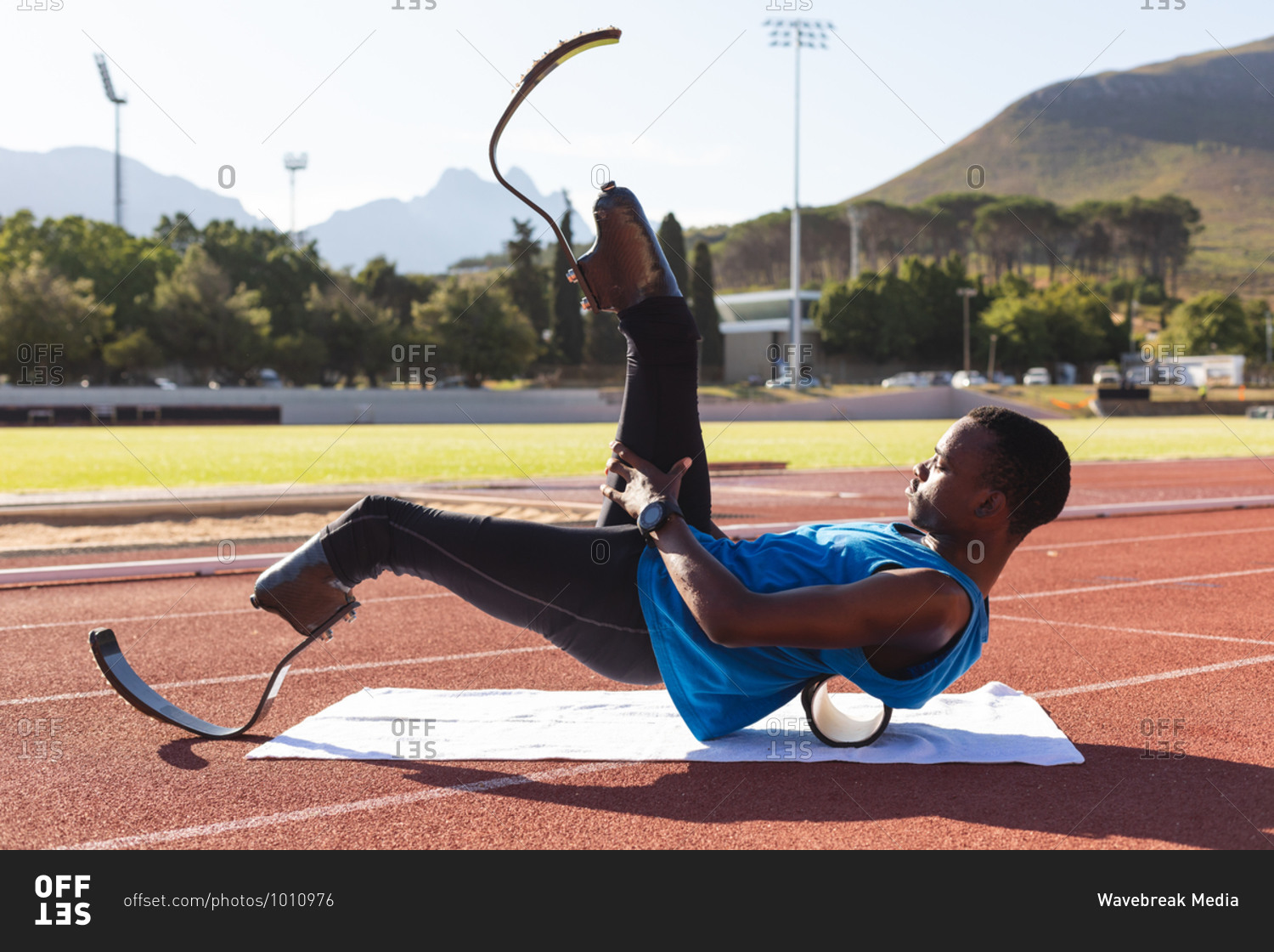 Fit, mixed race disabled male athlete at an outdoor sports stadium, preparing before workout stretching on roller on race track wearing running blades. Disability athletics sport training.