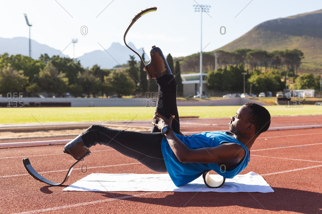Fit, mixed race disabled male athlete at an outdoor sports stadium, preparing before workout stretching on roller on race track wearing running blades. Disability athletics sport training.