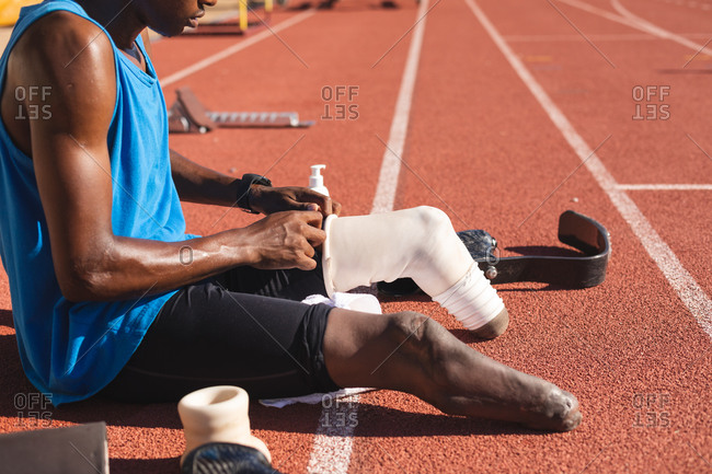 Mid section of fit, mixed race disabled male athlete at an outdoor sports stadium, sitting on race track preparing running blades before workout. Disability athletics sport training.