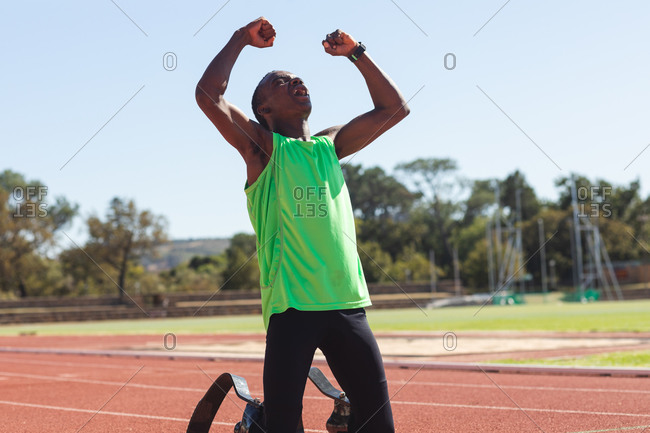 Fit, mixed race disabled male athlete at an outdoor sports stadium, kneeling on race track after race with arms in the air wearing running blades. Disability athletics sport training.