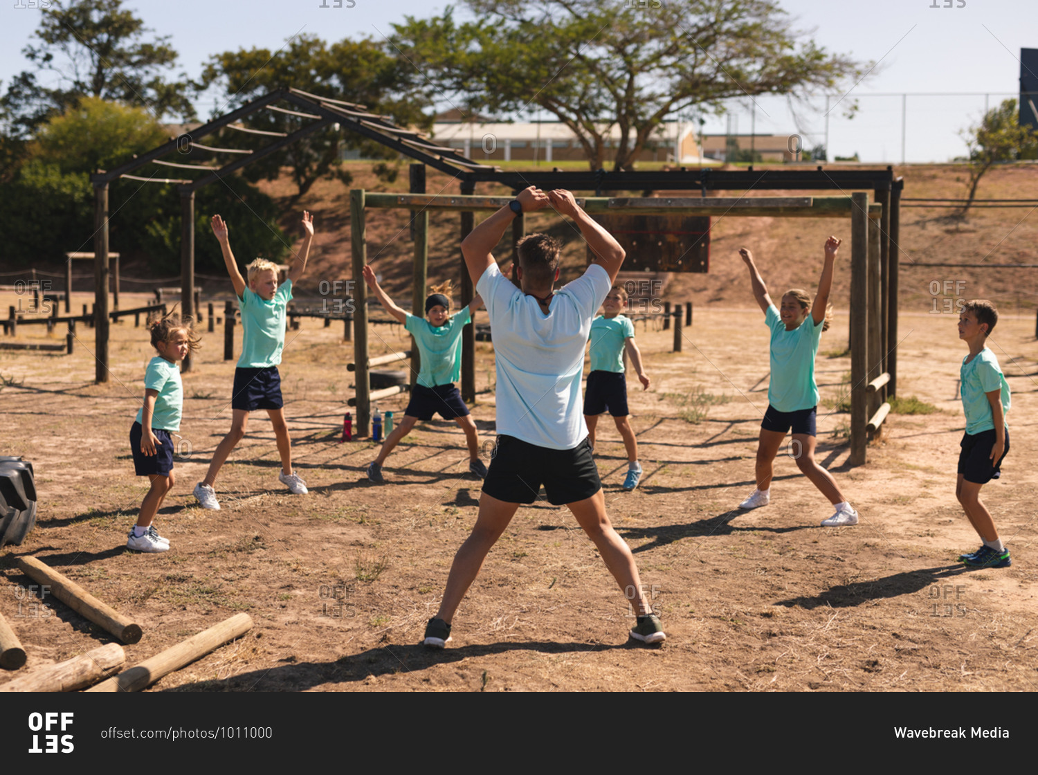 Rear view of Caucasian male fitness coach instructing a group of Caucasian boys girls at a boot camp on a sunny day doing jumping jacks wearing green t shirts and black shorts