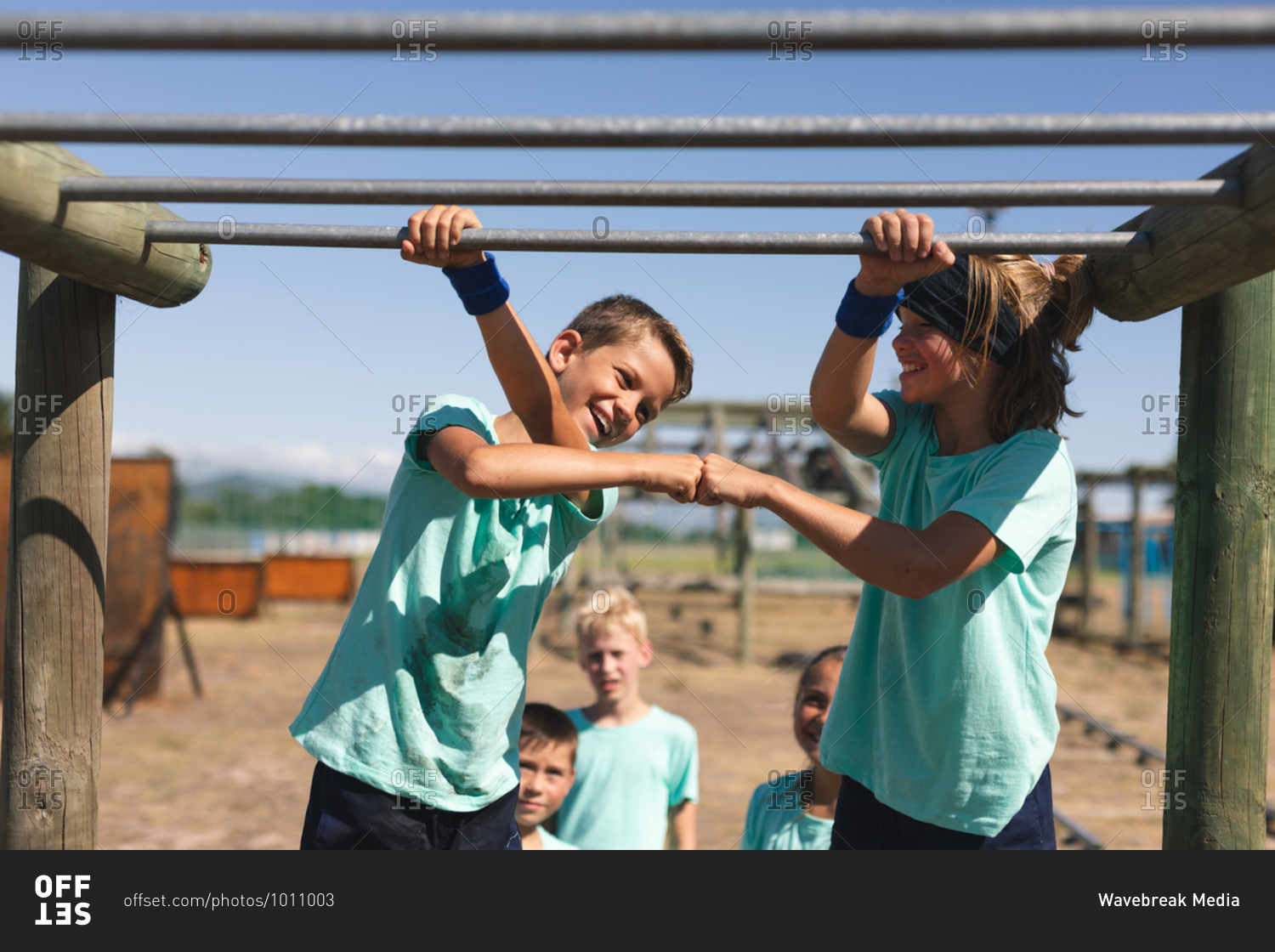 Two smiling Caucasian boys at a boot camp on a sunny day, standing beside each other holding on to the monkey bars, smiling and fist bumping, wearing green t shirts and black shorts