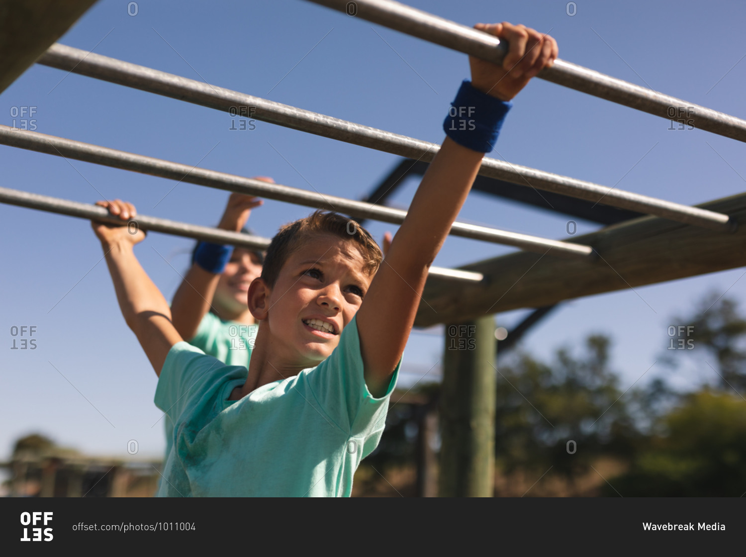Smiling Caucasian boy with brown hair at a boot camp on a sunny day, wearing green t shirt, on a jungle gym hanging from the monkey bars against a blue sky, another boy behind him in the background