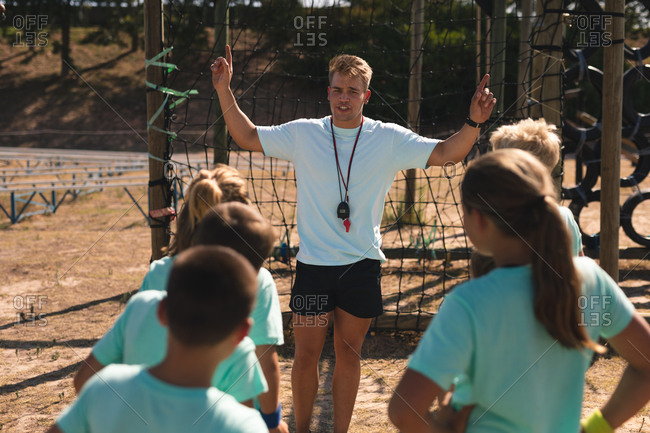 A group of Caucasian boys and girls listening to instructions from a Caucasian male fitness coach at a boot camp on a sunny day, standing and paying attention to him while he gestures and explains