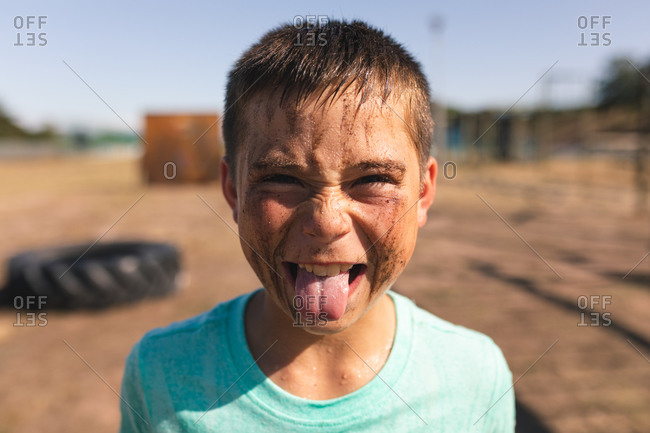 Portrait of Caucasian boy with short dark hair and mud on his face looking at camera, sticking his tongue out and making a face at a boot camp on a sunny day, wearing green t shirt
