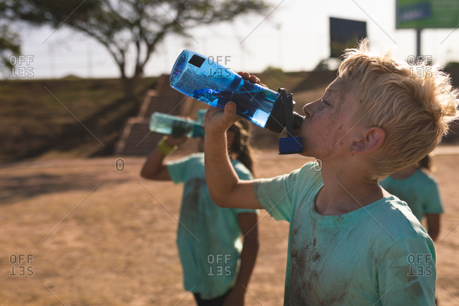A group of Caucasian boys and girls with muddy green t shirts and dirty faces having a rest and drinking bottles of water at a boot camp on a sunny day