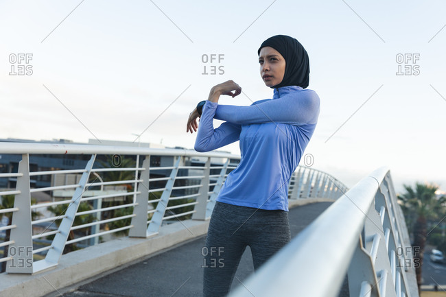 Fit mixed race woman  wearing hijab and sportswear exercising outdoors in the city on a sunny day, stretching her arms on a footbridge. Urban lifestyle exercise.