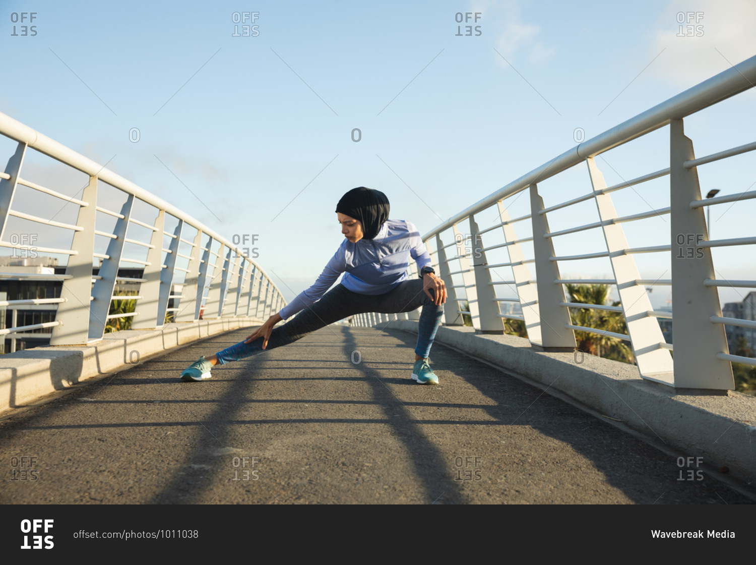 Fit mixed race woman wearing hijab and sportswear exercising outdoors in the city on a sunny day, stretching her legs on a footbridge. Urban lifestyle exercise.