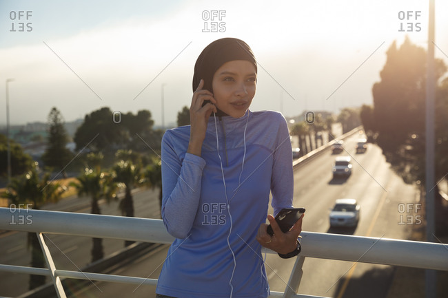 Fit mixed race woman wearing hijab and sportswear exercising outdoors in the city on a sunny day, taking break during workout using smartphone and earphones on a footbridge. Urban lifestyle exercise.