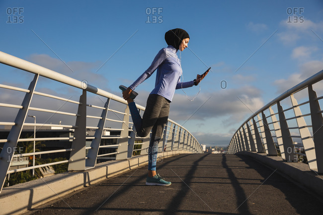 Fit mixed race woman wearing hijab and sportswear exercising outdoors in the city on a sunny day, stretching during workout using smartphone and earphones on a footbridge. Urban lifestyle exercise.
