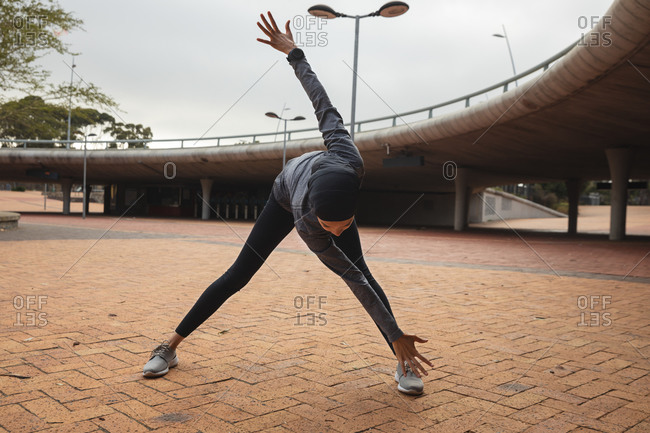Fit mixed race woman wearing hijab and sportswear exercising outdoors in the city, warming up stretching in urban park. Urban lifestyle exercise.