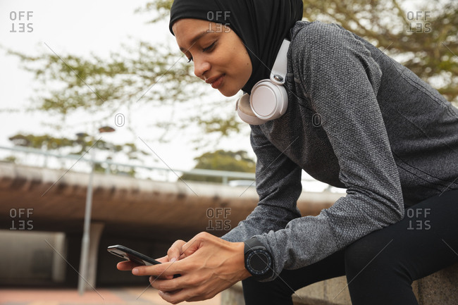 Fit mixed race woman wearing hijab and sportswear exercising outdoors in the city, sitting taking break using her smartphone in urban park. Urban lifestyle exercise.