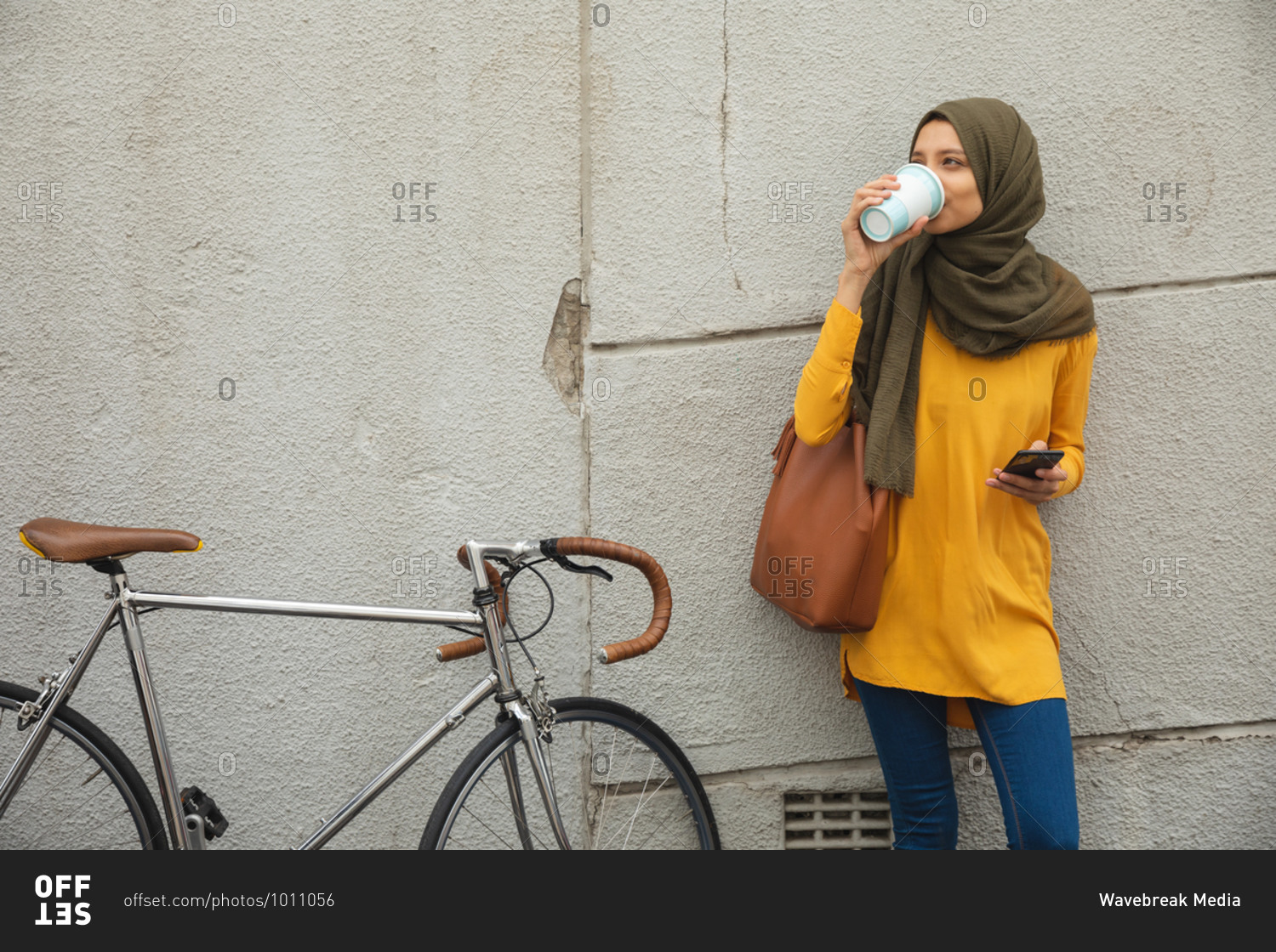 Mixed race woman wearing hijab and yellow jumper out and about in the city, standing by wall drinking takeaway coffee holding smartphone bike next to her. Commuter modern lifestyle.