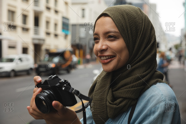 Mixed race woman wearing hijab sightseeing out and about in the city, smiling holding digital camera taking photos. Tourism sightseeing modern lifestyle.