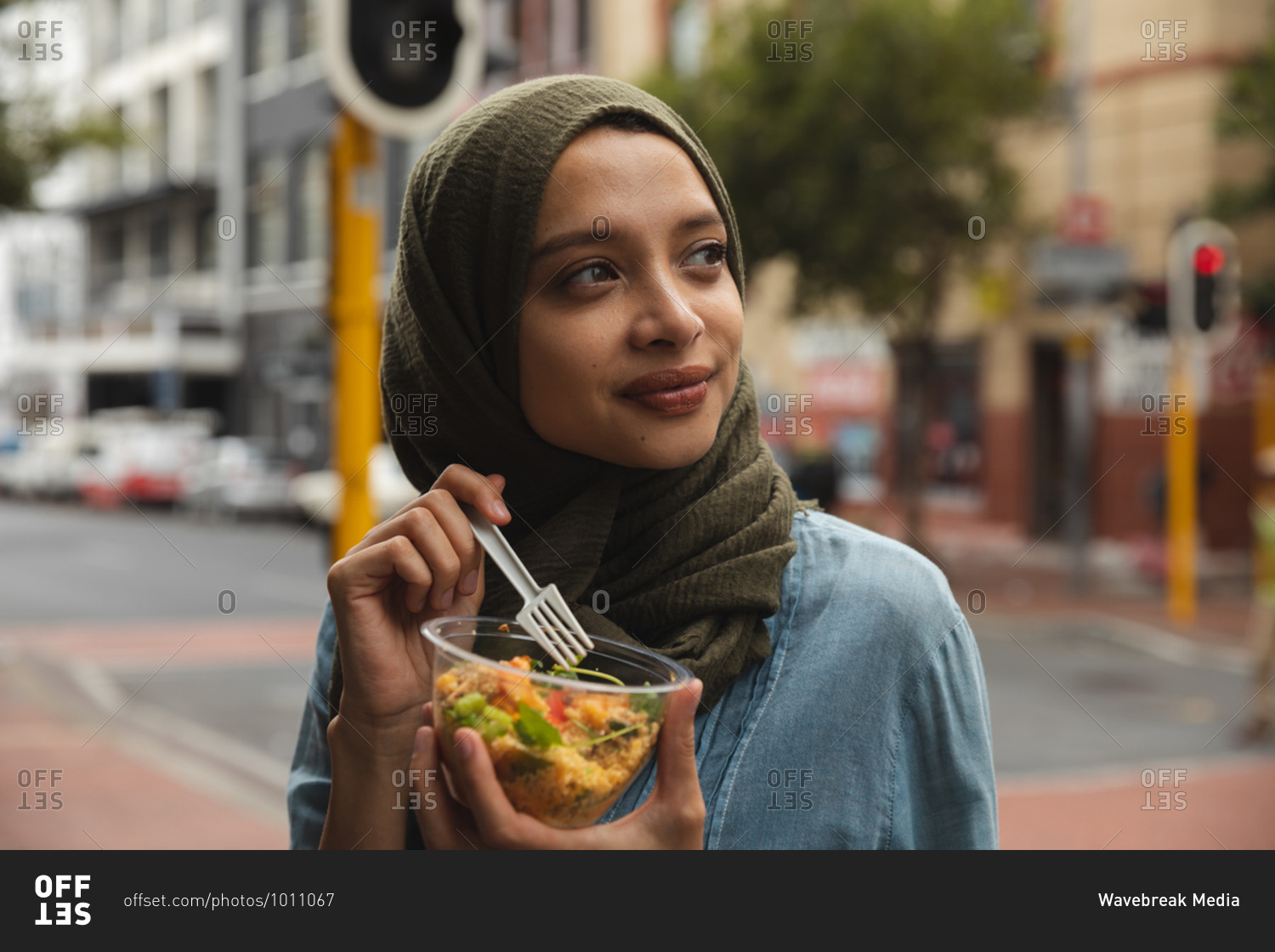 Mixed race woman wearing hijab out and about in the city, standing in street eating takeaway lunch holding bowl and fork, smiling. Commuter modern lifestyle.