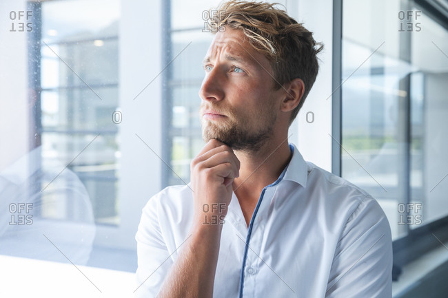 Smart casually dressed Caucasian male business creative with blonde hair and beard looking out of window thinking, with hand on his chin. Creative business professional working in a modern office.