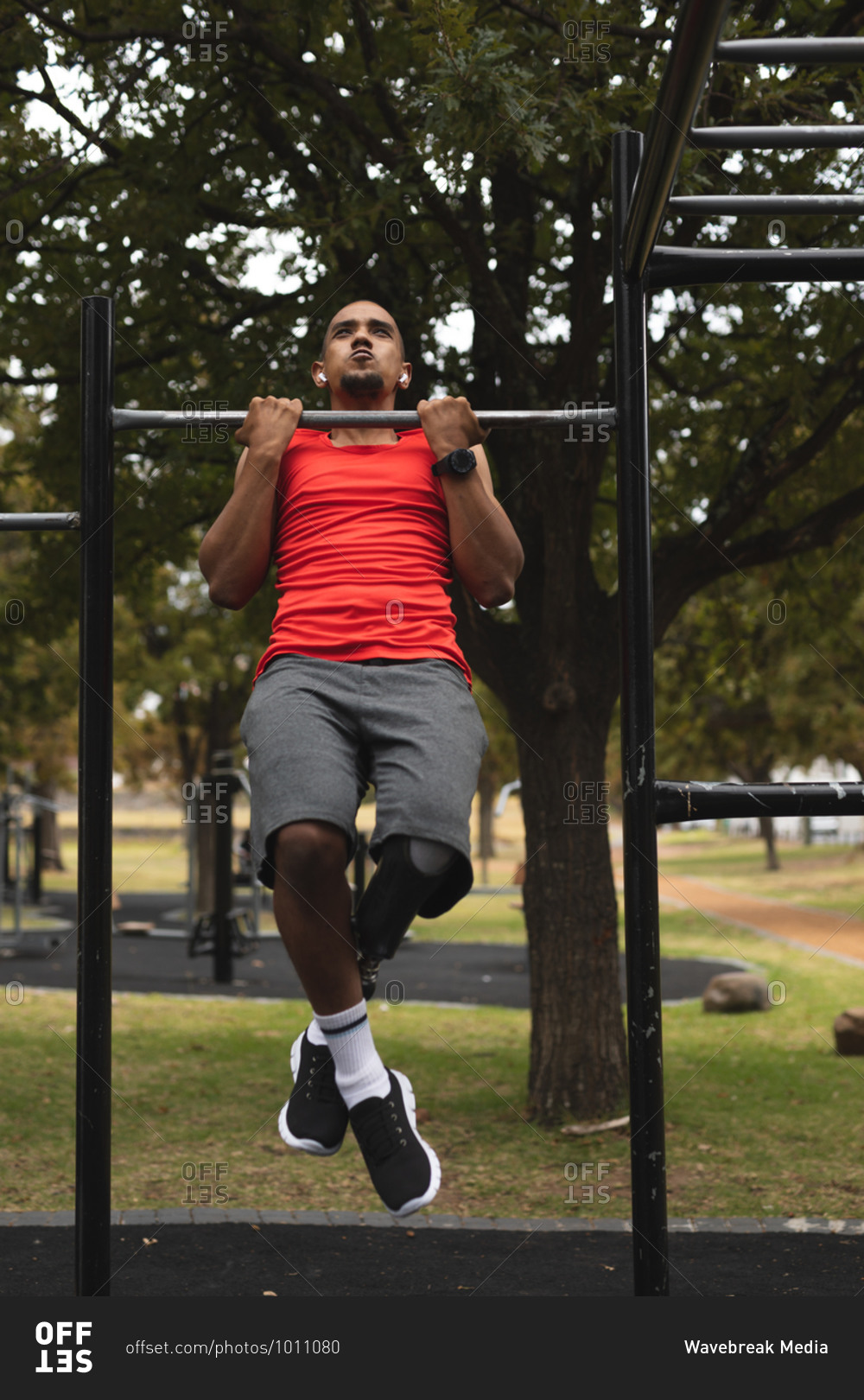 Disabled mixed race man with a prosthetic leg, working out in a park in outdoor gym, with wireless earphones on doing pull ups. Fitness disability healthy lifestyle.