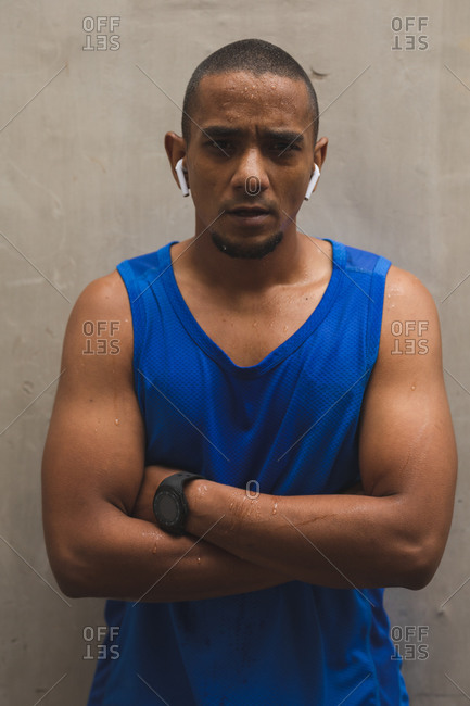 Portrait of mixed race man working out in an urban park, wearing sportswear, smartwatch and wireless earphones, taking a break, looking to camera with arms crossed. Fitness strength healthy lifestyle.
