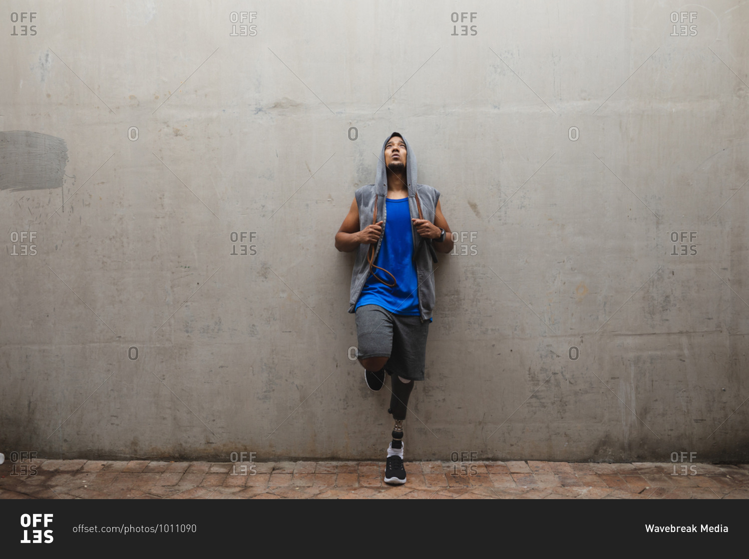Disabled mixed race man with a prosthetic leg, working out in an urban park, wearing hooded top leaning against a wall holding skipping rope taking a break. Fitness disability healthy lifestyle.