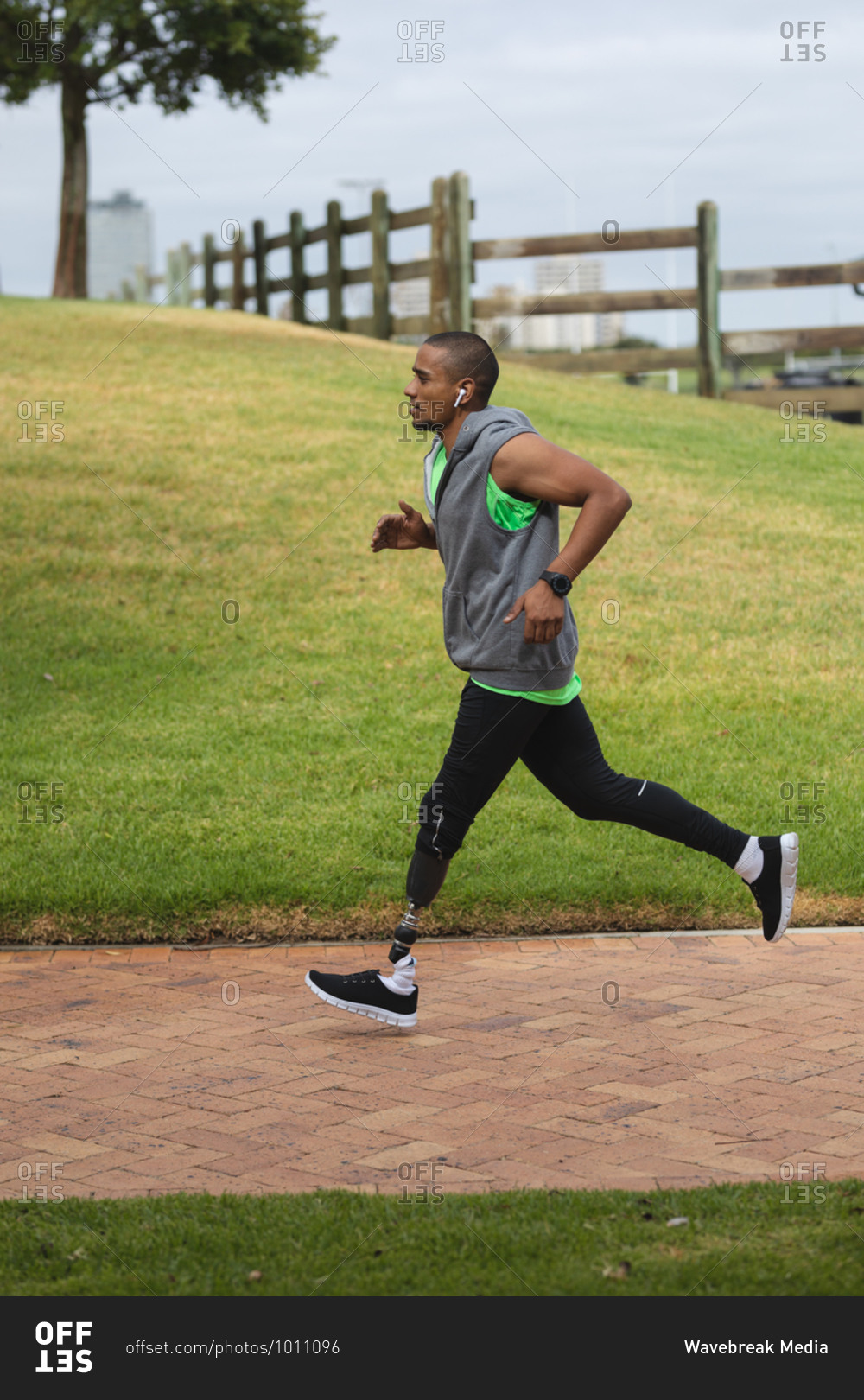 Disabled mixed race man with a prosthetic leg, working out in an urban park, wearing hooded top, running on a path. Fitness disability healthy lifestyle.