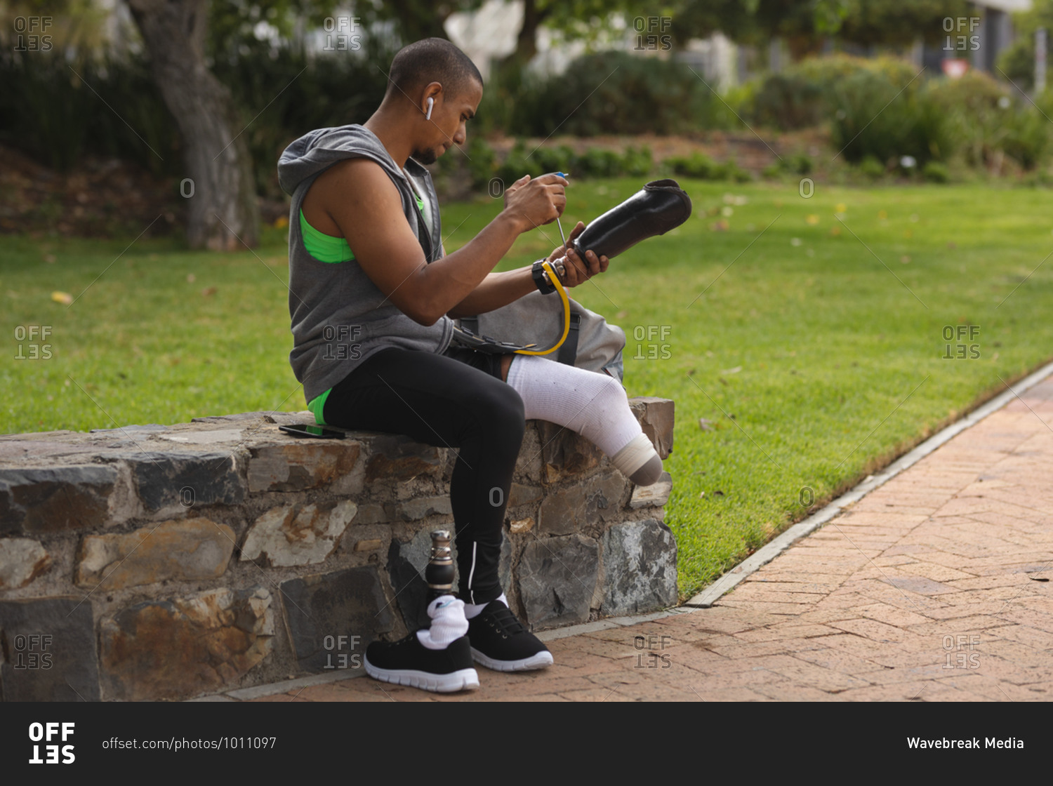 Disabled mixed race man with a prosthetic leg, working out in an urban park, sitting on a wall by a path making adjustments to his prosthetic leg. Fitness disability healthy lifestyle.