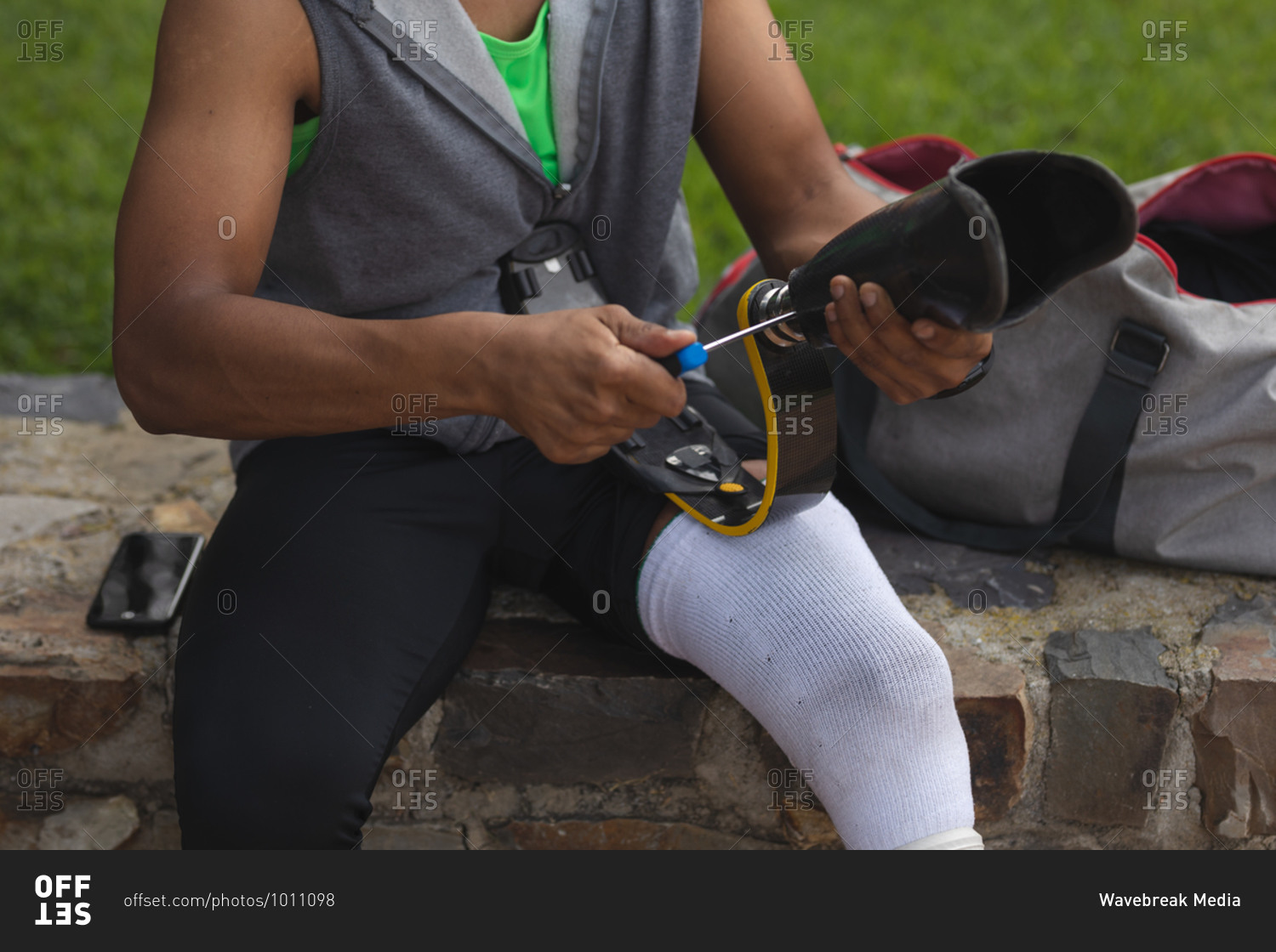 Mid section of a disabled mixed race man with a prosthetic leg working out in an urban park, sitting on a wall and fitting a running blade. Fitness disability healthy lifestyle.