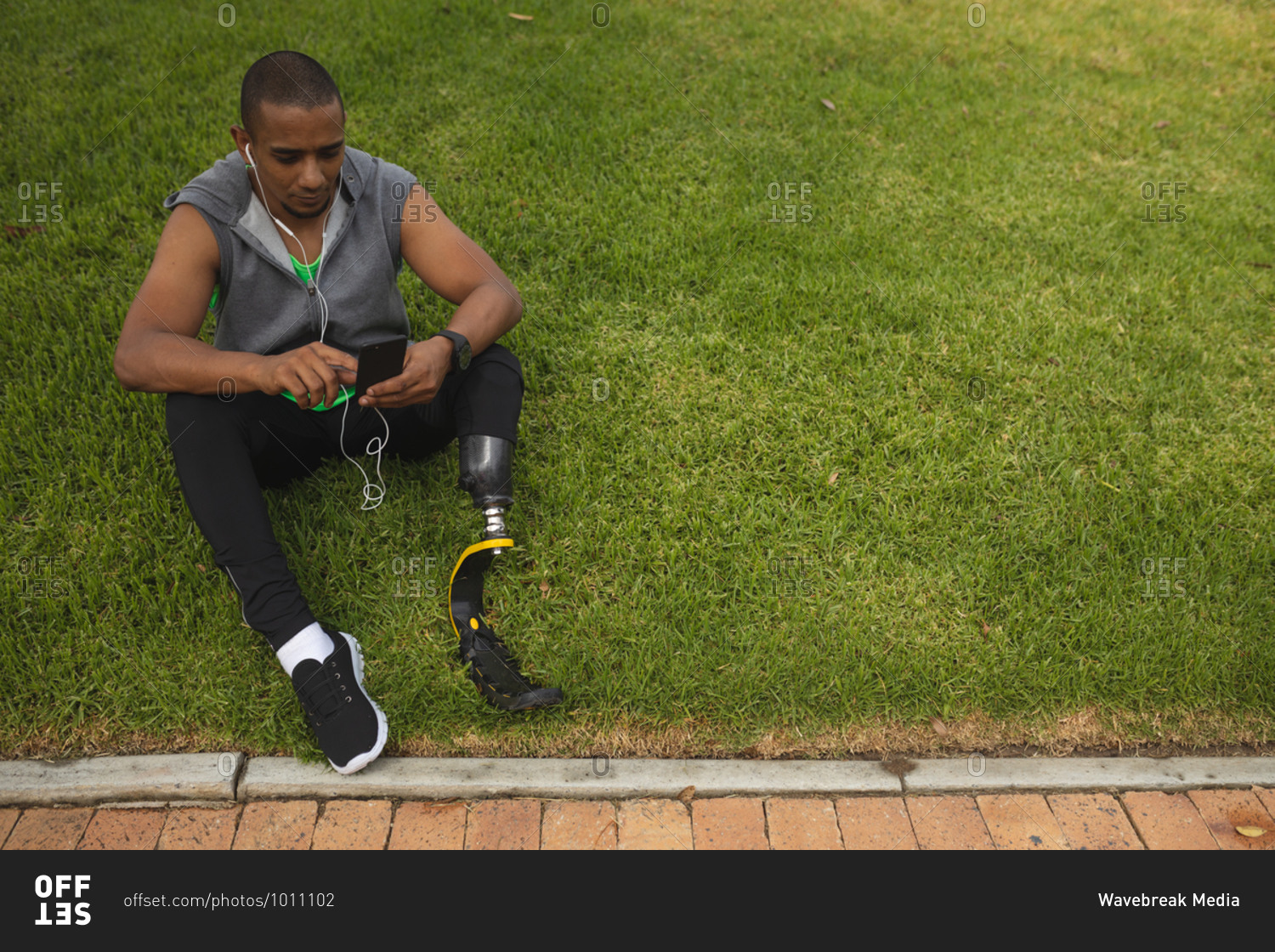 Disabled mixed race man with a prosthetic leg and running blade working out in a park, taking a break, sitting on grass using smartphone and wearing earphones. Fitness disability healthy lifestyle.