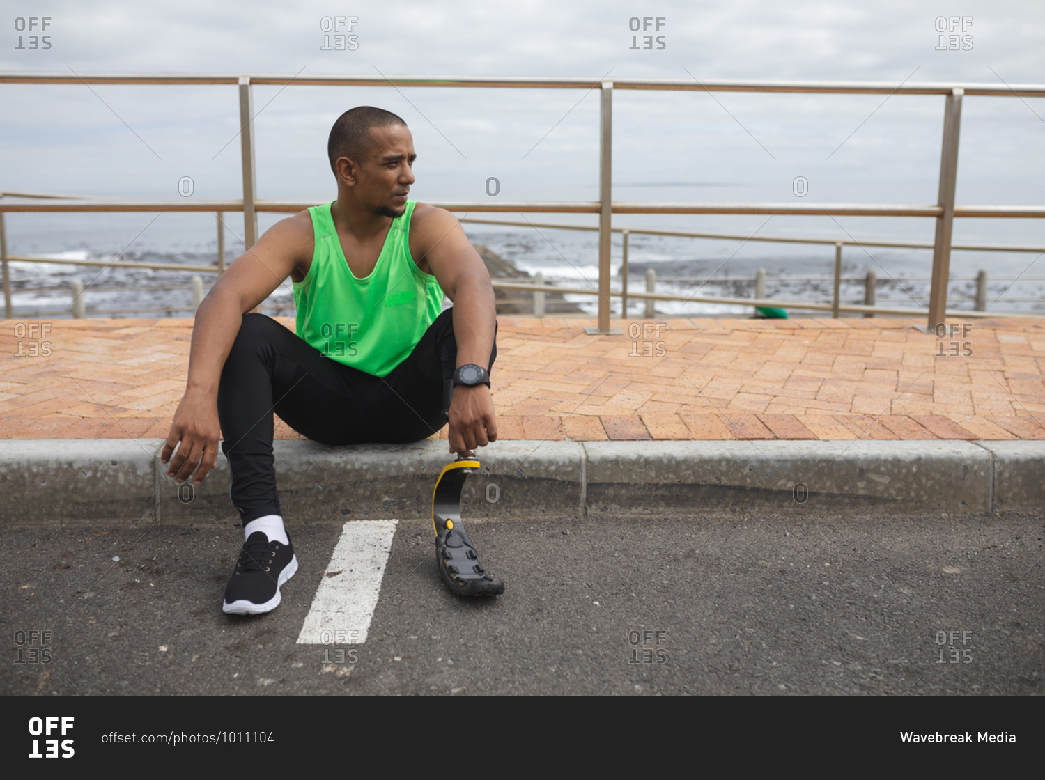 Disabled mixed race man with a prosthetic leg and running blade working out by the coast, sitting on the pavement by a road and taking a break. Fitness disability healthy lifestyle.