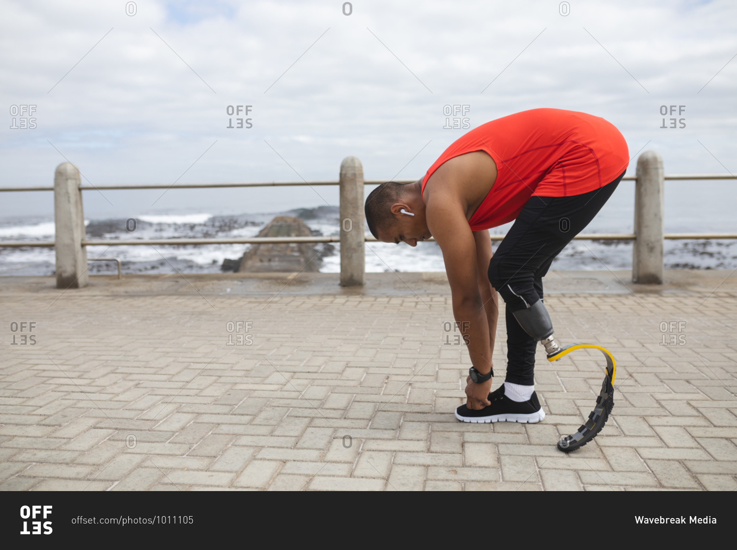 Disabled mixed race man with a prosthetic leg and running blade working out by the coast wearing wireless earphones, bending down to tie his shoelace. Fitness disability healthy lifestyle.