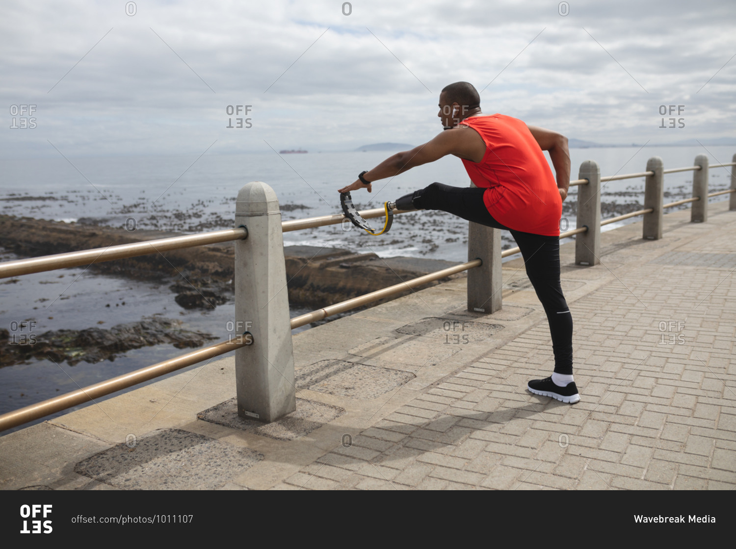 Disabled mixed race man with a prosthetic leg and running blade working out by the coast wearing wireless earphones, stretching legs with his blade on a fence. Fitness disability healthy lifestyle.