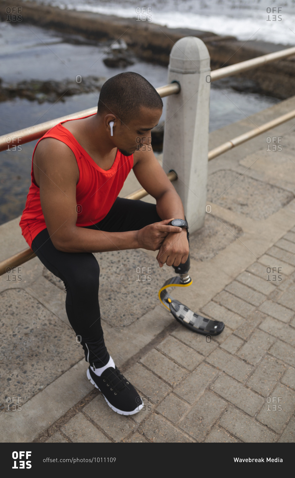 Disabled mixed race man with a prosthetic leg and running blade working out by the coast wearing wireless earphones, sitting on a fence and checking smartwatch. Fitness disability healthy lifestyle.