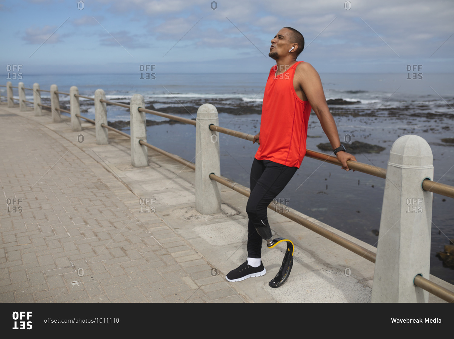 Disabled mixed race man with a prosthetic leg and running blade working out by the coast wearing wireless earphones, taking a break leaning on a fence. Fitness disability healthy lifestyle.