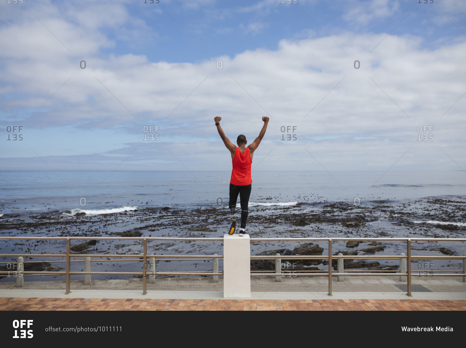 Rear view of disabled mixed race man with a prosthetic leg and running blade working out by the coast, standing on a fence looking out to sea with arms raised. Fitness disability healthy lifestyle.