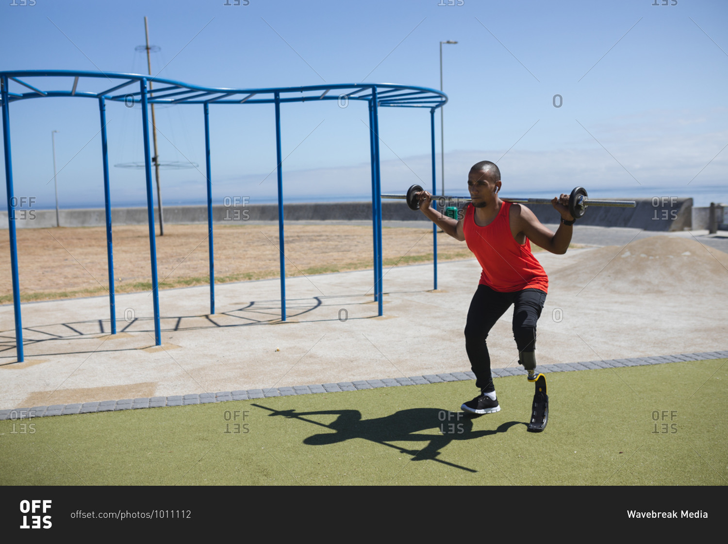 Disabled mixed race man with a prosthetic leg and running blade working out at an outdoor gym by the coast, lifting weights on a barbell. Fitness disability healthy lifestyle.