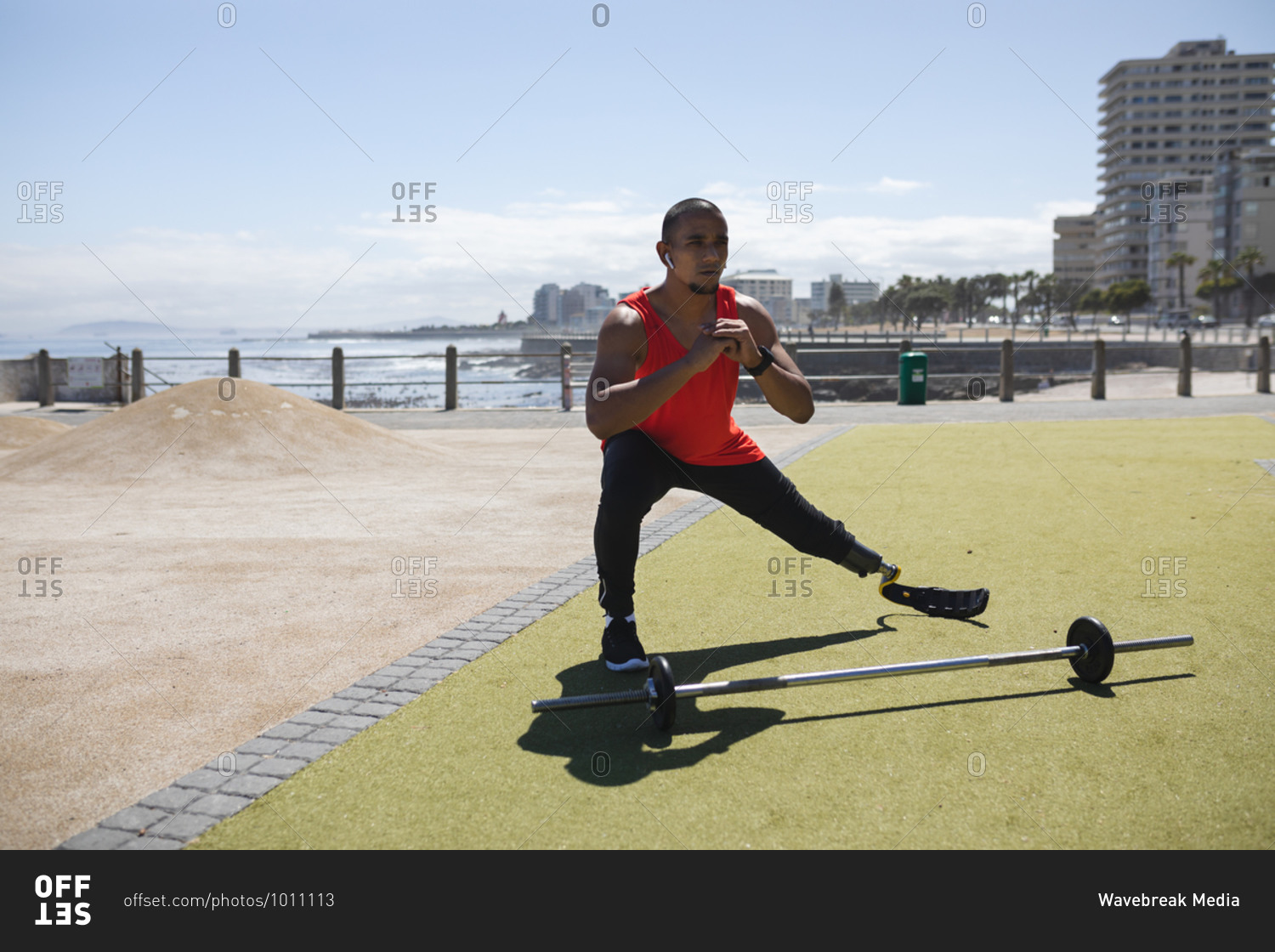 Disabled mixed race man with a prosthetic leg and running blade at an outdoor gym by the coast wearing wireless earphones, stretching legs, a barbell beside him. Fitness disability healthy lifestyle.