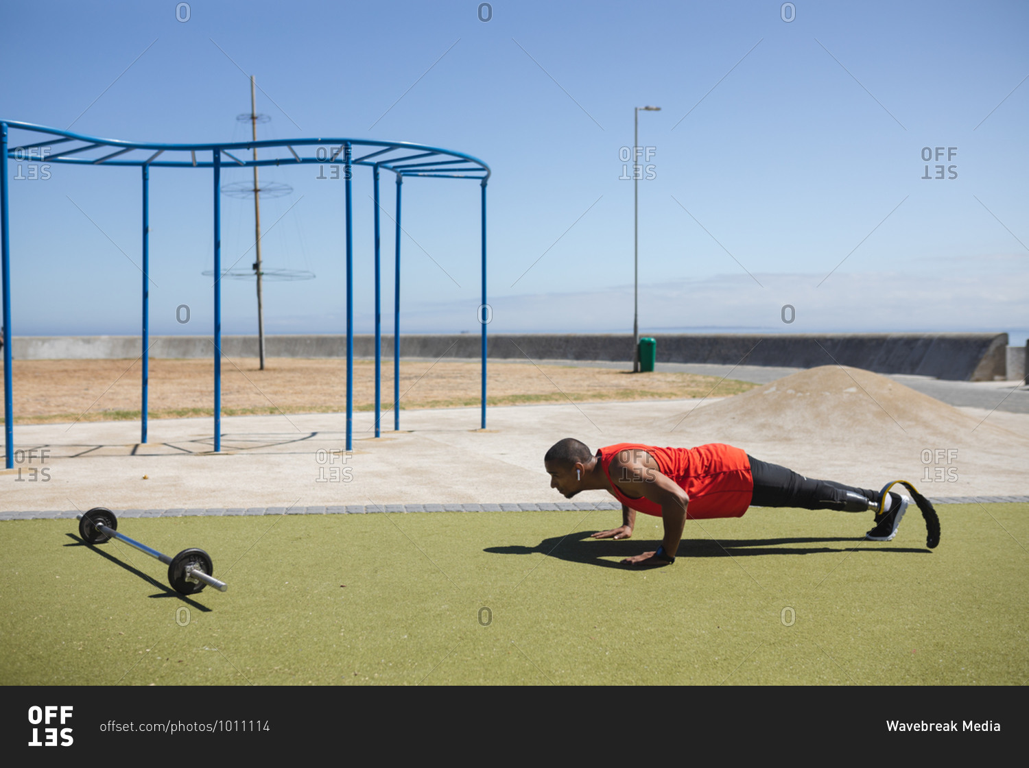 Disabled mixed race man with a prosthetic leg and running blade at an outdoor gym by the coast wearing wireless earphones, doing press ups, a barbell beside him. Fitness disability healthy lifestyle.