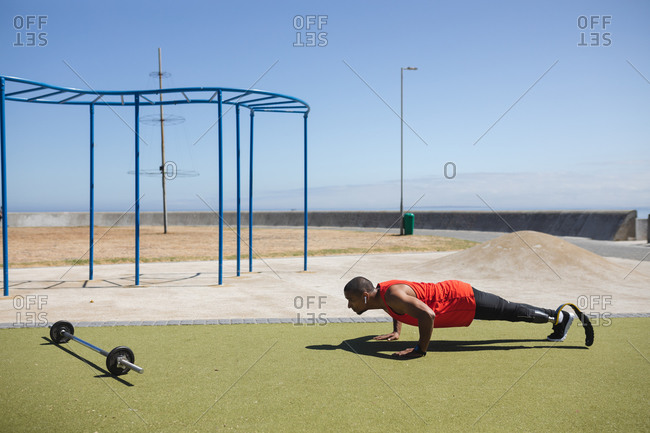 Disabled mixed race man with a prosthetic leg and running blade at an outdoor gym by the coast wearing wireless earphones, doing press ups, a barbell beside him. Fitness disability healthy lifestyle.