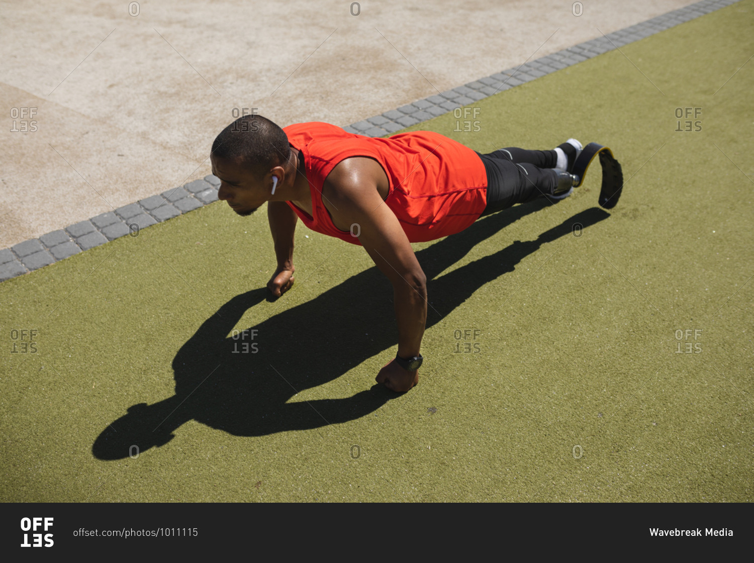 Disabled mixed race man with a prosthetic leg and running blade exercising at an outdoor gym by the coast wearing wireless earphones and doing press ups. Fitness disability healthy lifestyle.