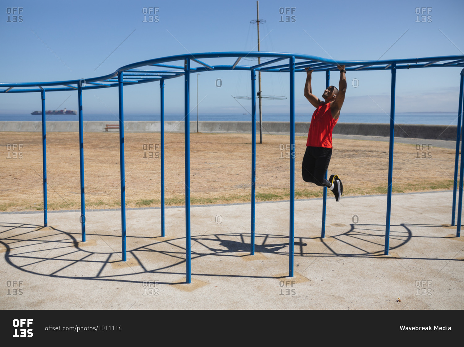 Disabled mixed race man with a prosthetic leg and running blade exercising at an outdoor gym by the coast, working out on the monkey bars. Fitness disability healthy lifestyle.