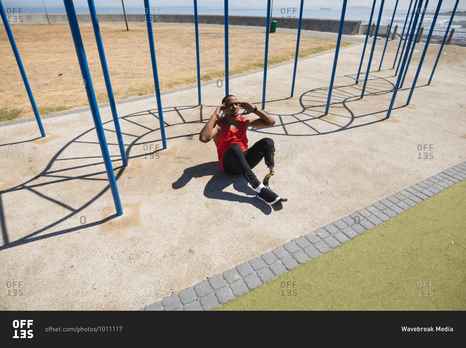 Disabled mixed race man with a prosthetic leg and running blade exercising at an outdoor gym by the coast, doing sit ups in the sun beside the gym equipment. Fitness disability healthy lifestyle.