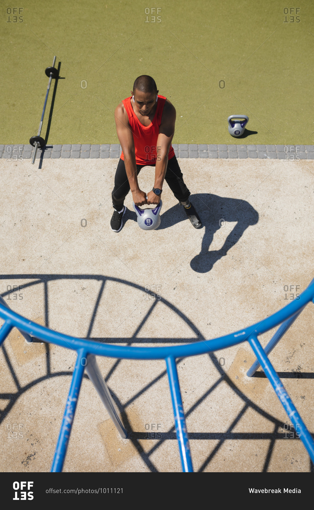 High angle view of disabled mixed race man with a prosthetic running blade working out at an outdoor gym wearing wireless earphones, swinging a kettlebell weight. Fitness disability healthy lifestyle.